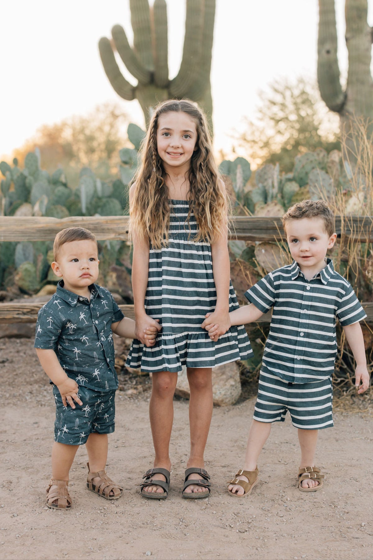 Three children wearing striped outfits stand in front of cacti, smiling and holding hands, in a desert setting.