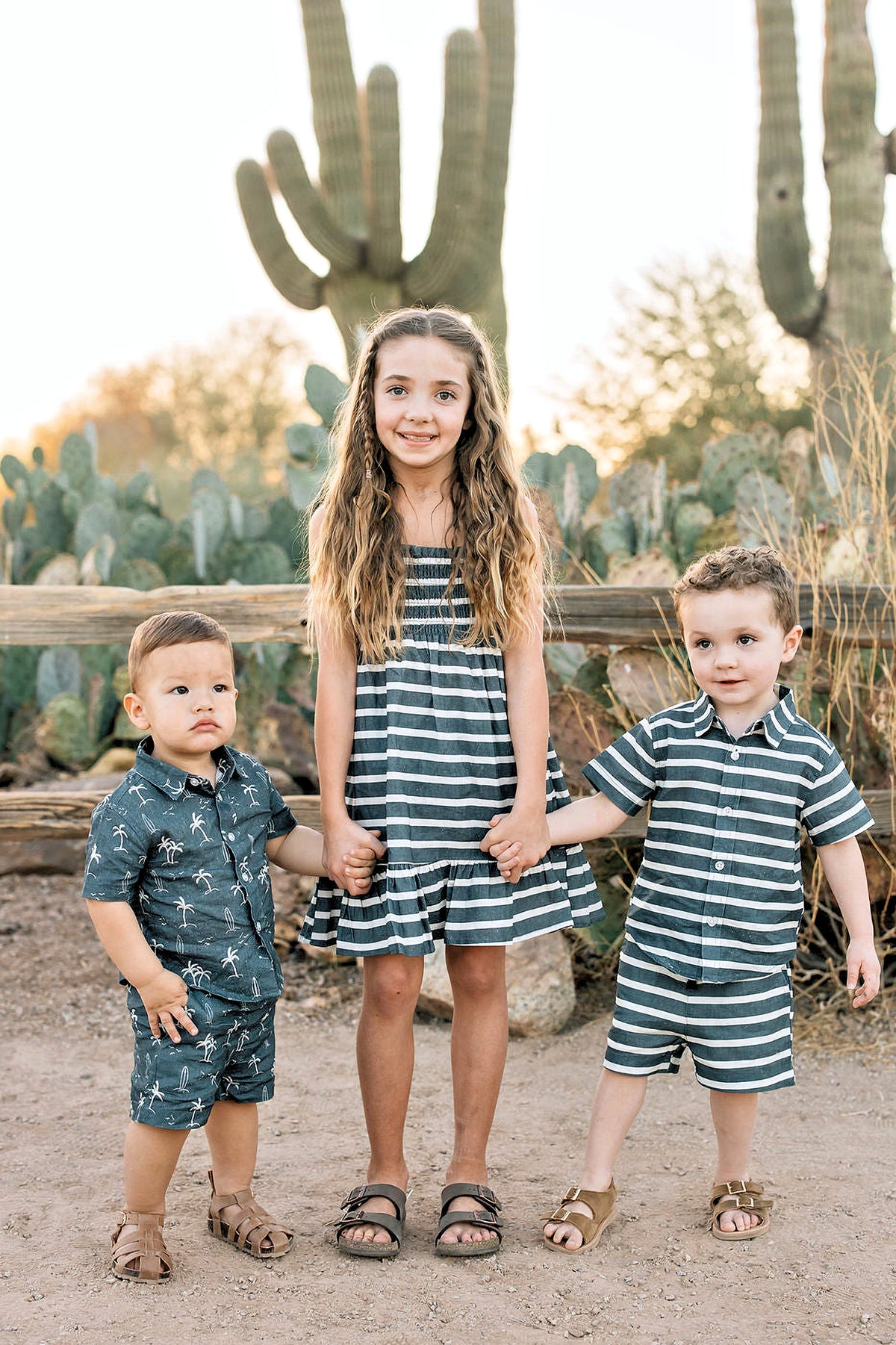 Three children standing in front of cacti, wearing striped and printed outfits, holding hands, and smiling.