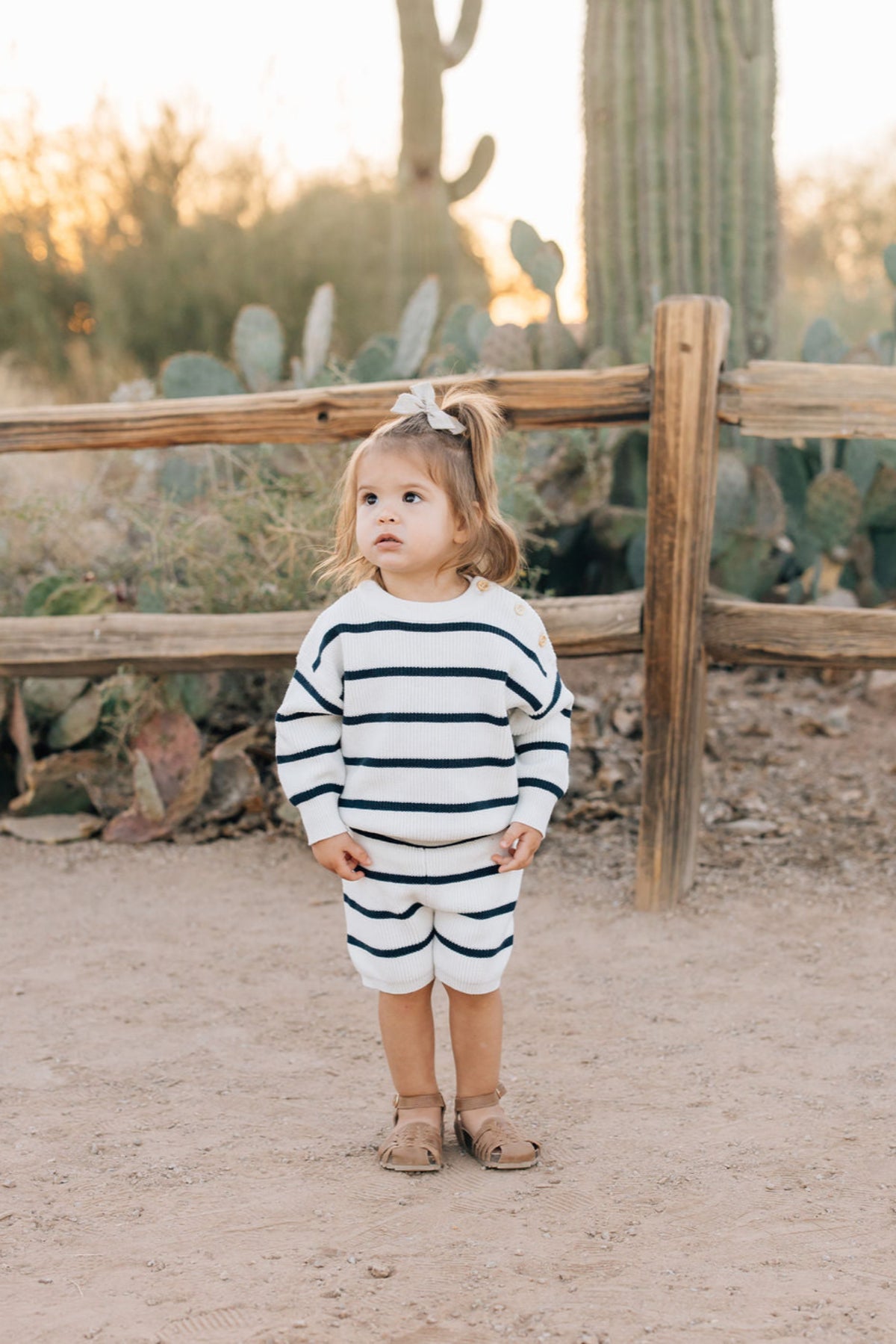 A toddler wearing a navy and white striped knit outfit stands outdoors near a wooden fence and cactus.
