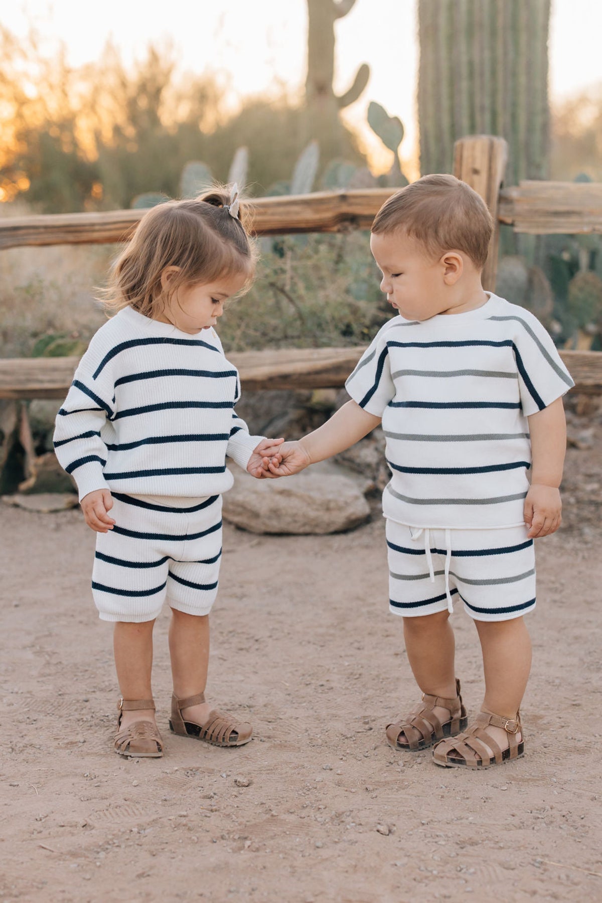 Two toddlers wearing matching navy and white striped knit outfits hold hands outdoors near a wooden fence.