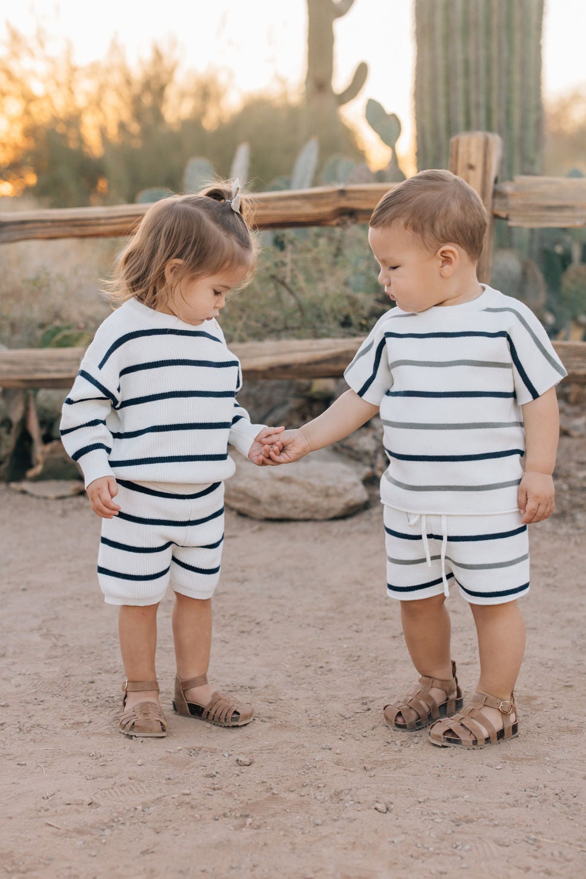 Two toddlers wearing matching striped knit outfits hold hands outdoors, with a wooden fence and cacti background.