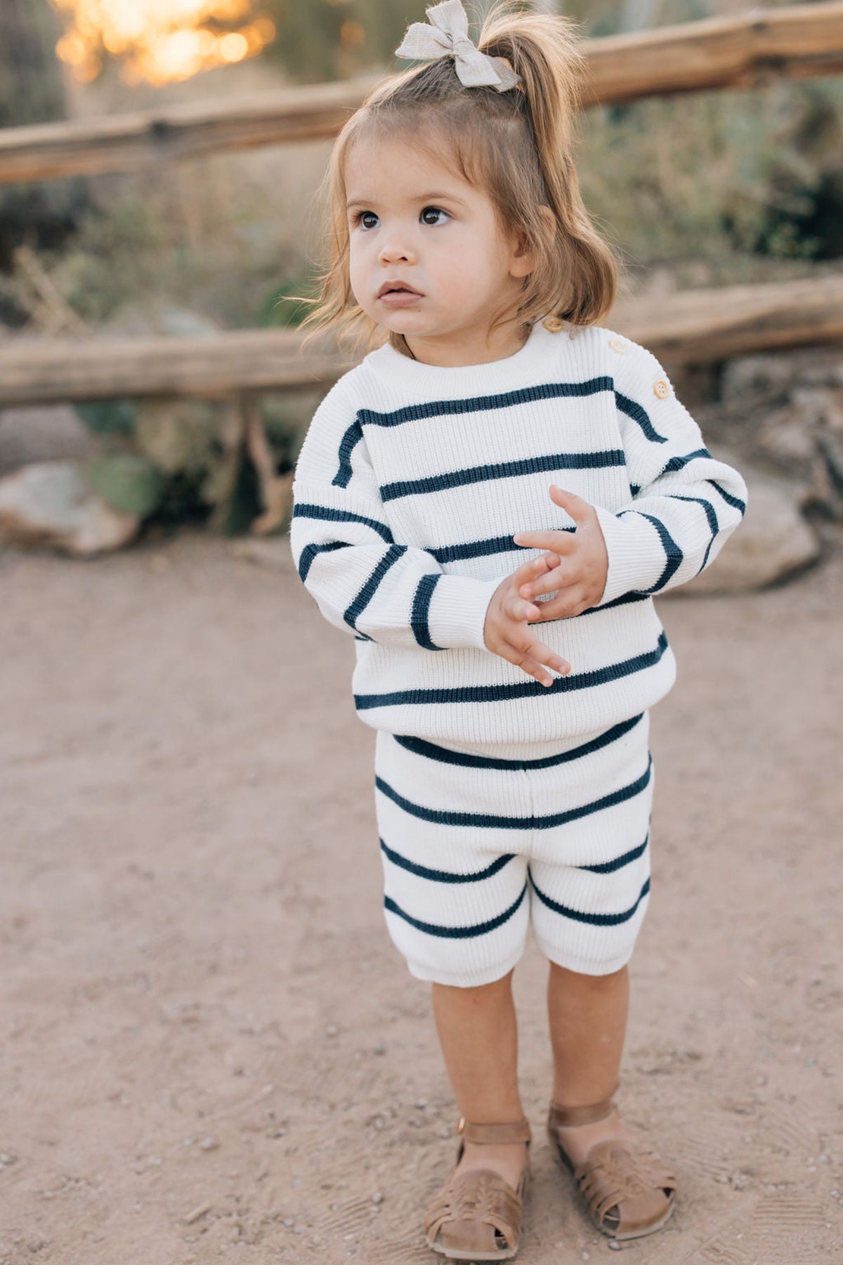 A toddler wearing a navy-striped knit outfit stands outdoors, with a bow in their hair and brown sandals.