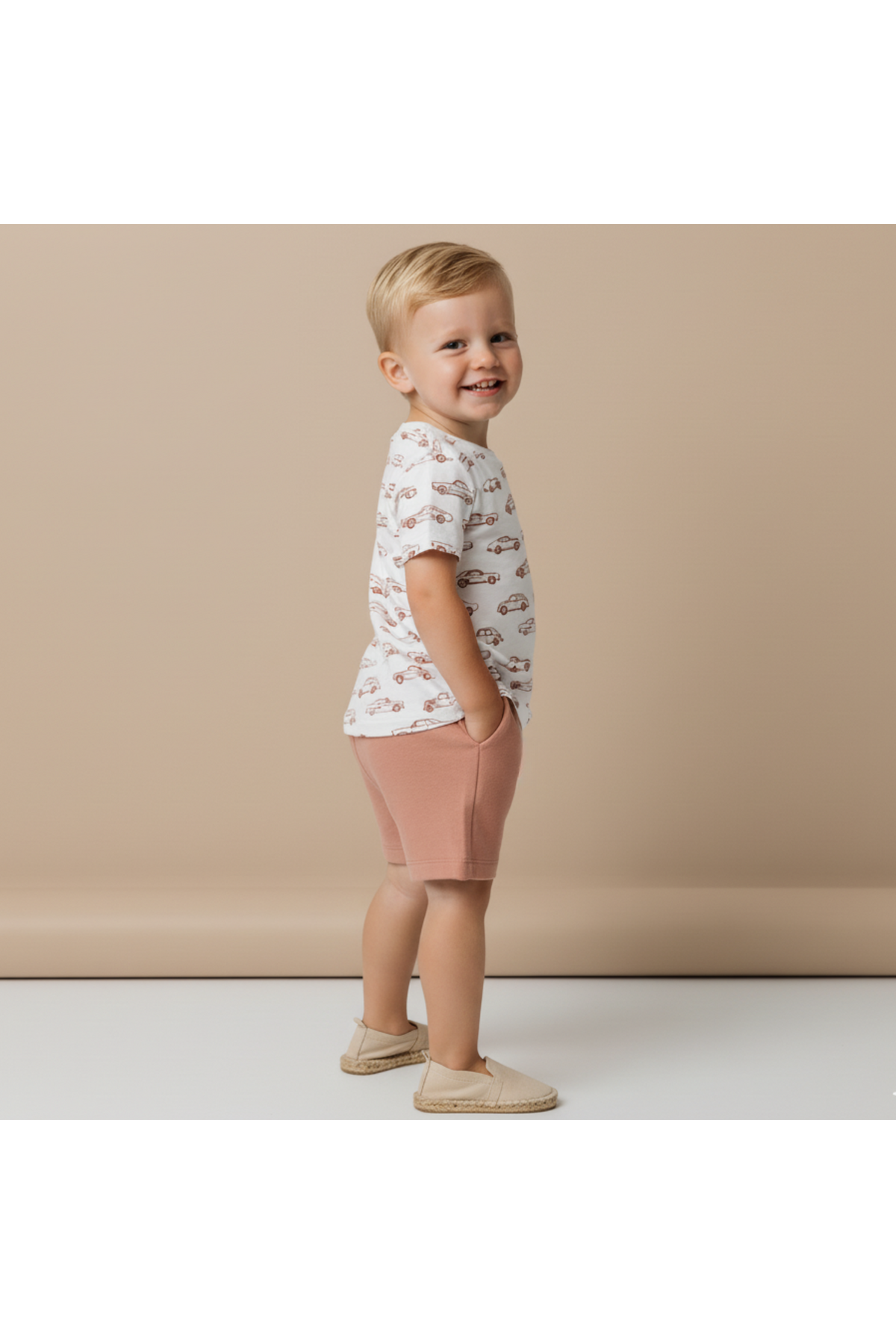 A smiling child stands sideways, wearing a white organic cotton tee with a playful car print and light pink shorts. The background is a soft beige, enhancing the outfits charm.