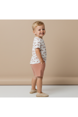 A smiling child stands sideways, wearing a white organic cotton tee with a playful car print and light pink shorts. The background is a soft beige, enhancing the outfits charm.