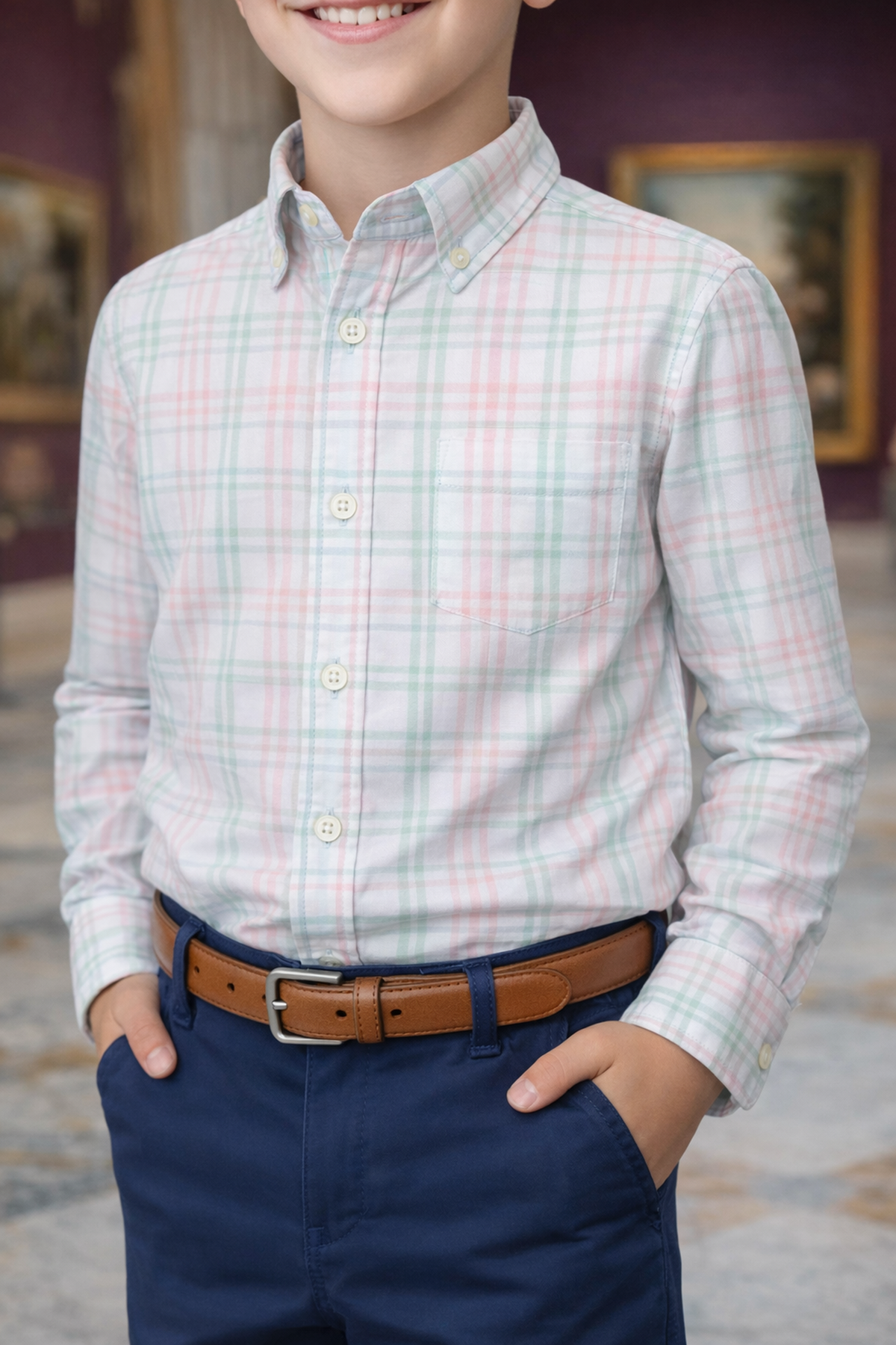 A young boy smiles while wearing a light plaid button-down shirt and navy shorts, complemented by a brown belt.