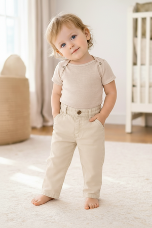A toddler stands on a soft rug, wearing a beige short-sleeve shirt and light-colored pants. The room features natural light, enhancing the cozy, family-friendly atmosphere.