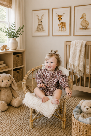 A smiling baby girl sits in a bamboo chair, wearing a vintage floral long sleeve romper.