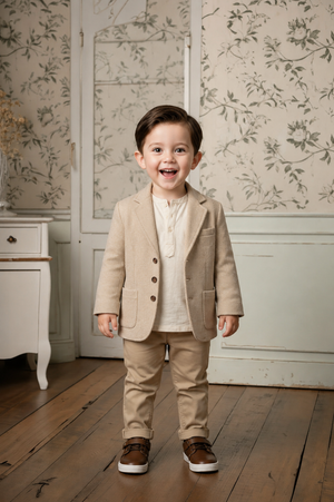 A cheerful boy stands in a stylish taupe blazer and matching trousers, smiling widely in a vintage-inspired room with floral wallpaper.