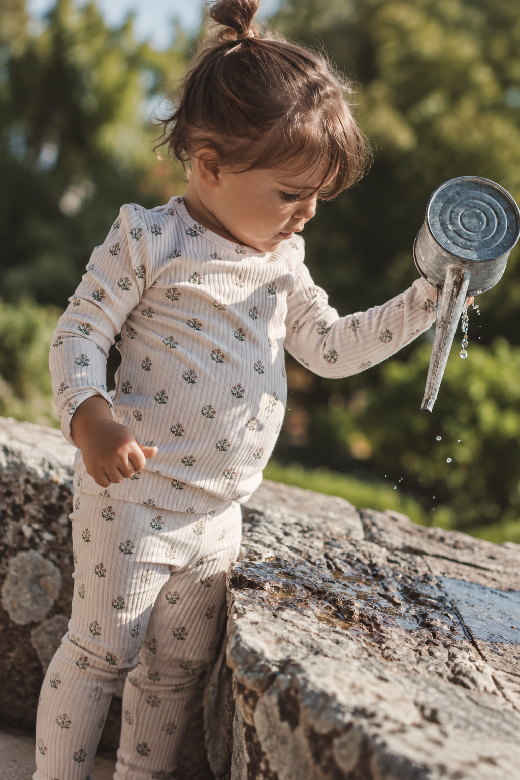 A toddler in a light-colored, ribbed outfit with small floral prints pours water from a metal watering can, standing on a stone surface surrounded by greenery.