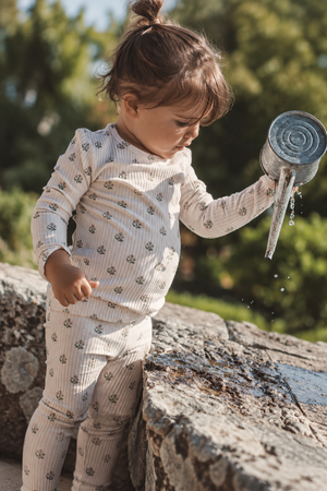 A toddler in a light-colored, ribbed outfit with small floral prints pours water from a metal watering can, standing on a stone surface surrounded by greenery.