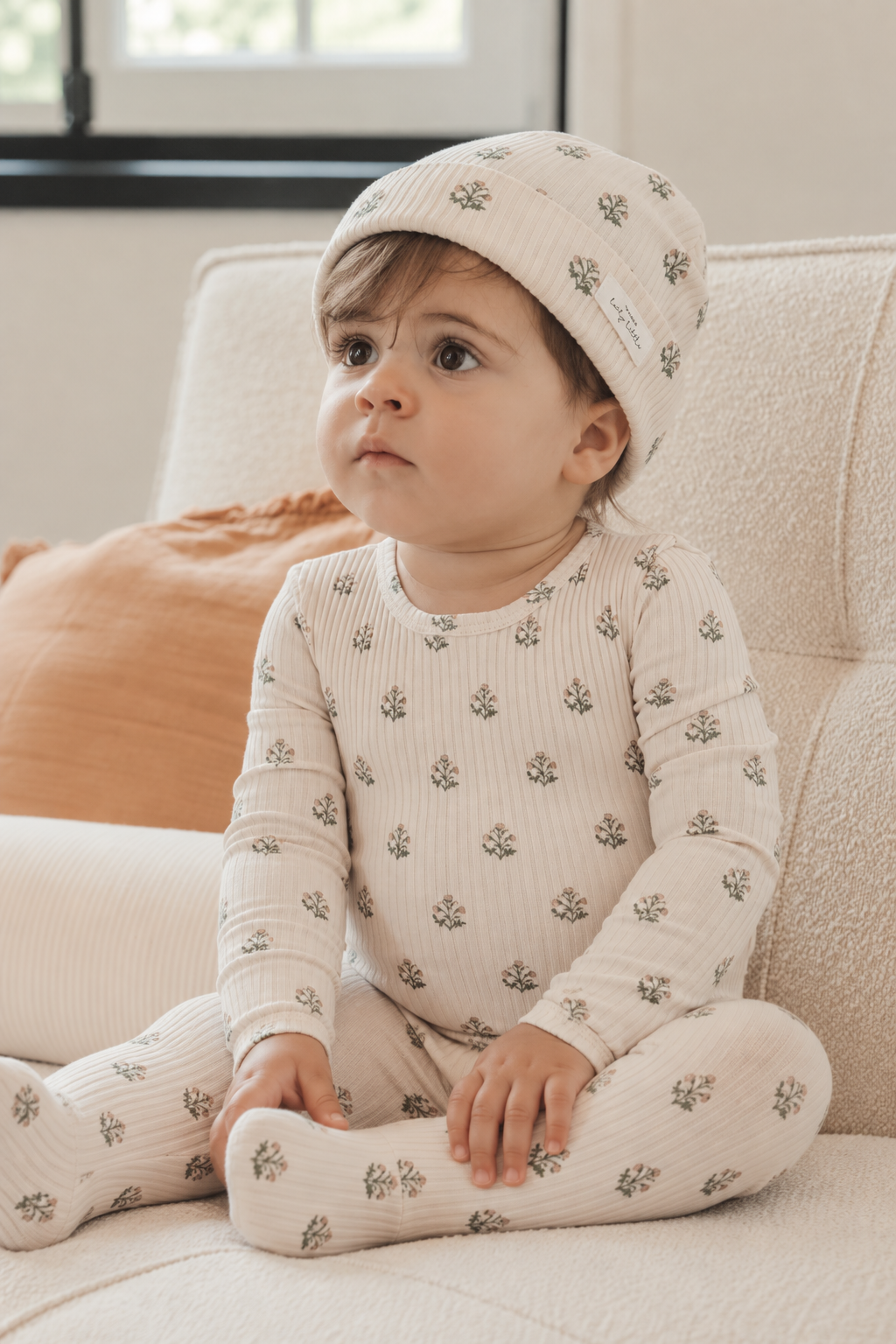 A young child sits on a textured cream sofa, wearing a light beige outfit with small floral prints and a matching hat.