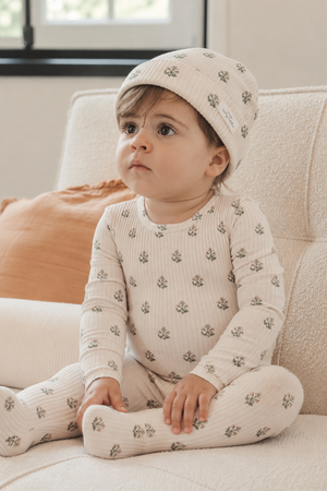 A young child sits on a textured cream sofa, wearing a light beige outfit with small floral prints and a matching hat.