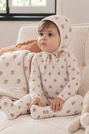 A young child sits on a cozy sofa, wearing a light beige outfit adorned with small floral patterns and a matching bonnet.