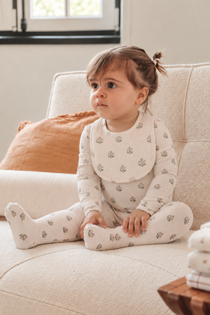 A toddler sits on a cozy, textured sofa, wearing a light-colored outfit with a floral print and a matching bib. The warm, inviting room features soft pillows.