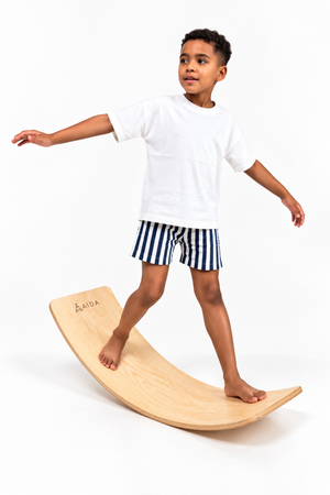 A young boy balances on a curved wooden balance board, wearing a white t-shirt and striped shorts. The board features a smooth finish, encouraging active and imaginative play.