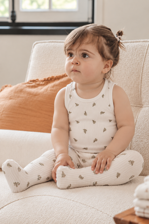 A toddler sits on a cozy sofa, wearing a white ribbed tank top featuring small green leaf patterns. The soft fabric adds comfort, perfect for warm summer days.