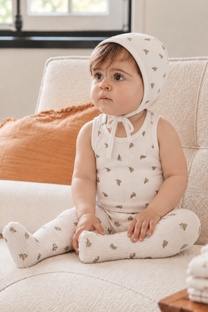 A young child sits on a light-colored sofa, wearing a white bonnet with a leaf print and a matching sleeveless outfit. The cozy setting features warm.