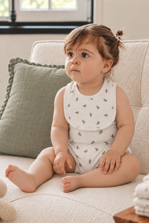 A toddler sits comfortably on a light-colored sofa, wearing a white cotton romper with a subtle leaf pattern. Soft pillows and natural light create a cozy atmosphere.
