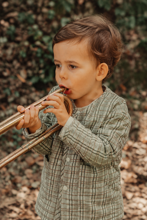 A young child plays a brass trombone while wearing a green plaid jacket. The setting features natural foliage, creating a vibrant, outdoor atmosphere.