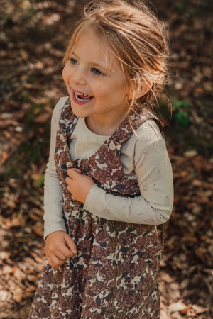 A smiling young girl wearing a floral corduroy dress stands in a wooded area. The dress features adjustable straps and is styled with a light long-sleeve shirt underneath.