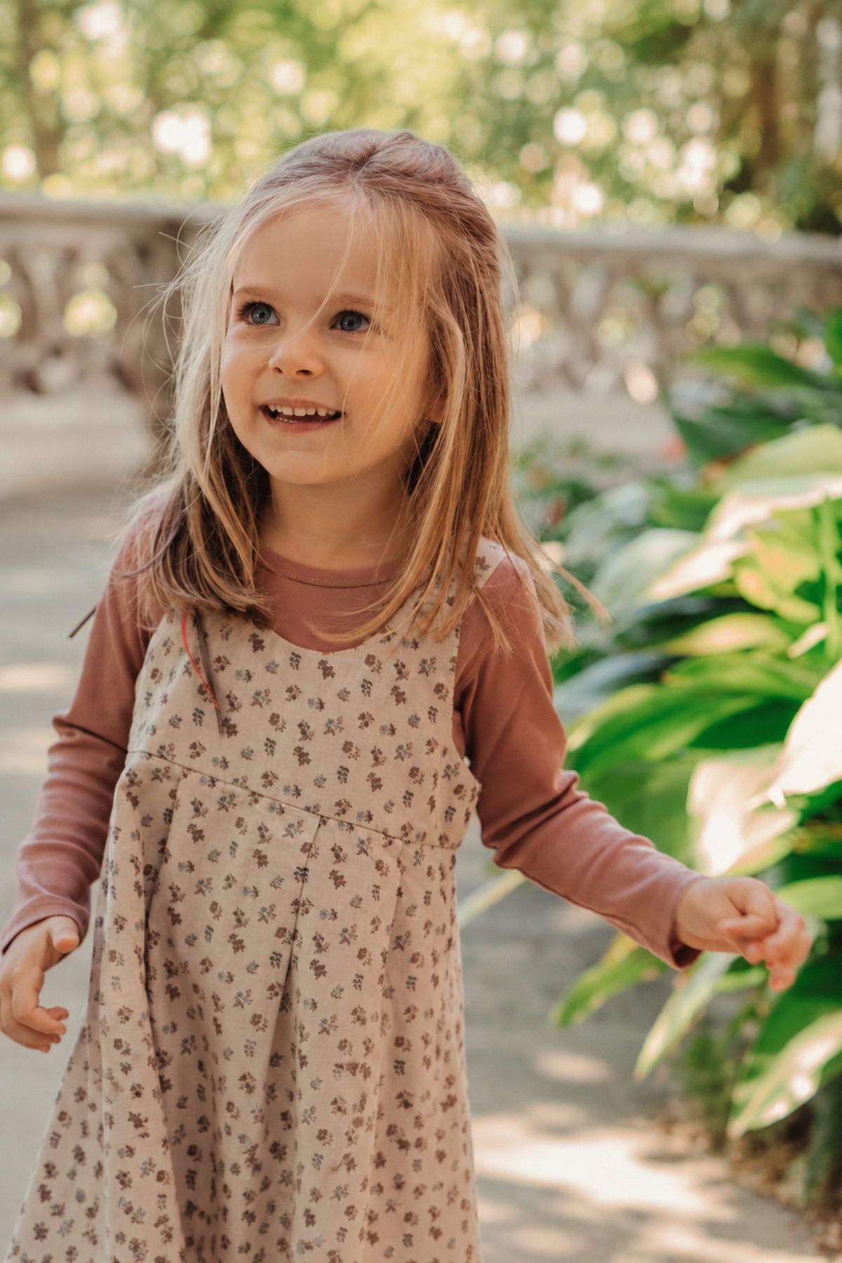A smiling young girl wearing a floral corduroy dress over a long-sleeve shirt stands amidst lush greenery, showcasing a charming and playful look perfect for outdoor activities.