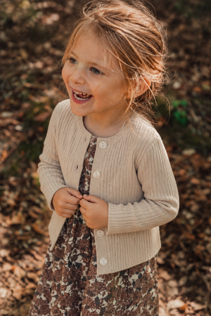 A joyful young girl wears a cream ribbed cotton cardigan over a floral dress, surrounded by autumn leaves. The cardigan features a button-up design, perfect for layering in cooler weather.