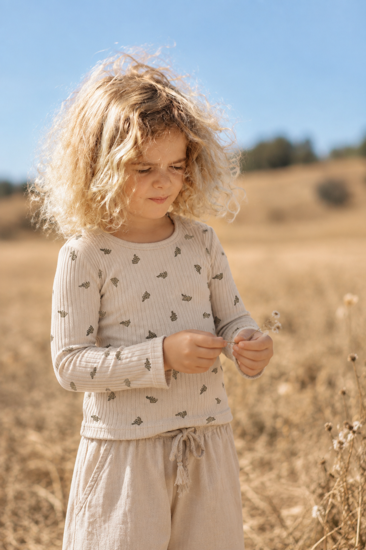 A young child with curly hair stands in a sunlit field, wearing a light beige long-sleeve ribbed tee adorned with small leaf patterns, paired with soft, light-colored pants.