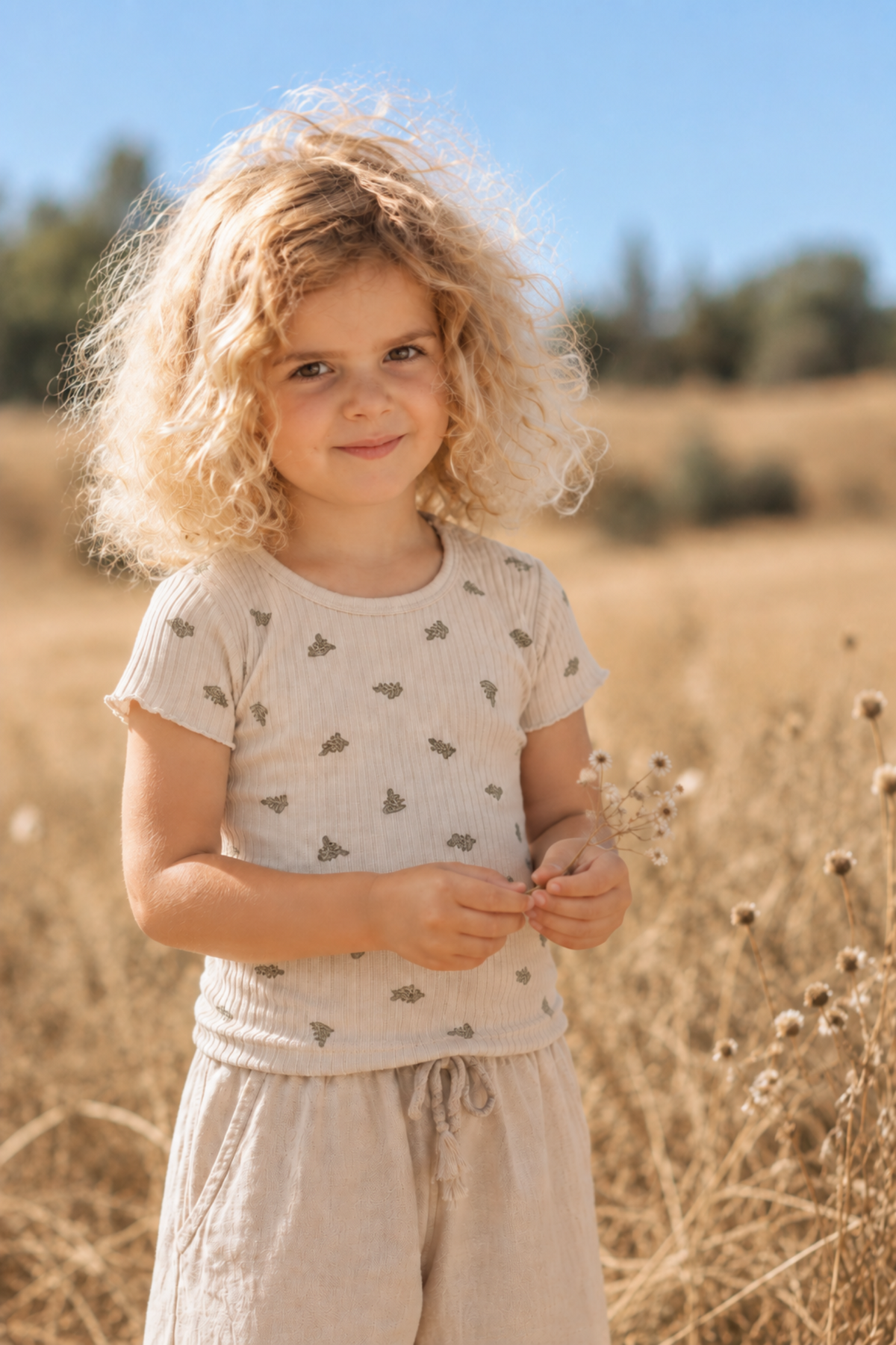 A young child stands in a field of dry grass, wearing a light beige short-sleeve tee with a subtle leaf print.