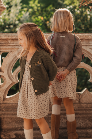 Two young girls are outdoors, wearing floral dresses and chunky knit cardigans. The girl in front sports an olive green cardigan with wooden buttons, showcasing a playful and cozy style.