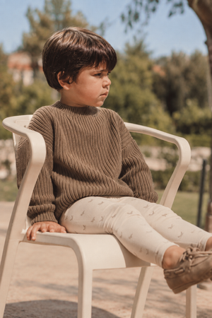 A young child sits on a white chair outdoors, wearing a chunky knit brown sweater and light-colored leggings with a subtle pattern.