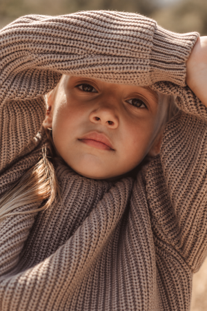 A close-up of a child wearing a chunky knit sweater in a warm beige color, showcasing a relaxed fit and textured fabric.