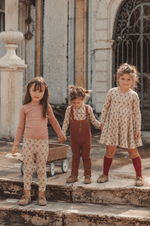 Three children stand together outdoors, dressed in coordinated outfits featuring soft, earthy colors.