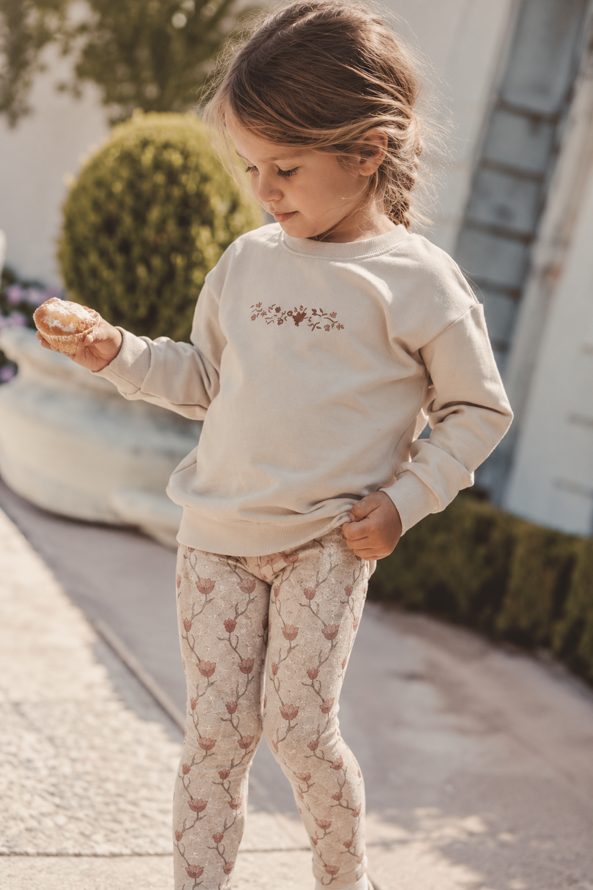 A young girl stands outside, wearing a beige sweatshirt with floral embroidery and light pink patterned leggings. She holds a pastry, showcasing a casual, playful outfit perfect for kids.