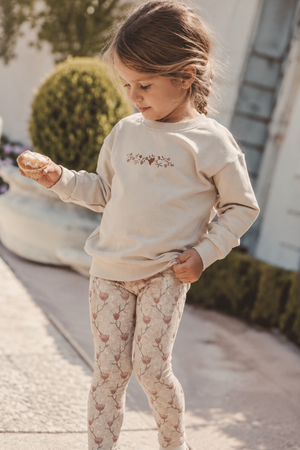 A young girl stands outside, wearing a beige sweatshirt with floral embroidery and light pink patterned leggings. She holds a pastry, showcasing a casual, playful outfit perfect for kids.