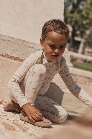 A young child sits on a stone surface, wearing a light beige turtleneck and matching pants adorned with a subtle horse print. The setting features natural outdoor elements.