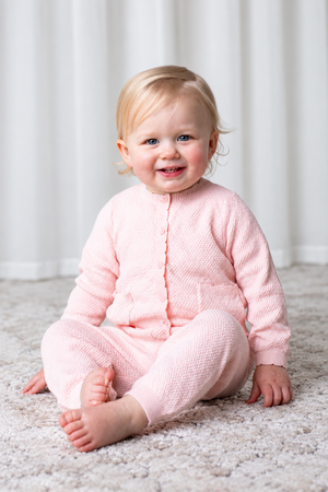 A smiling toddler sits on a soft, light-colored carpet, wearing a pale pink organic cotton sweater set.