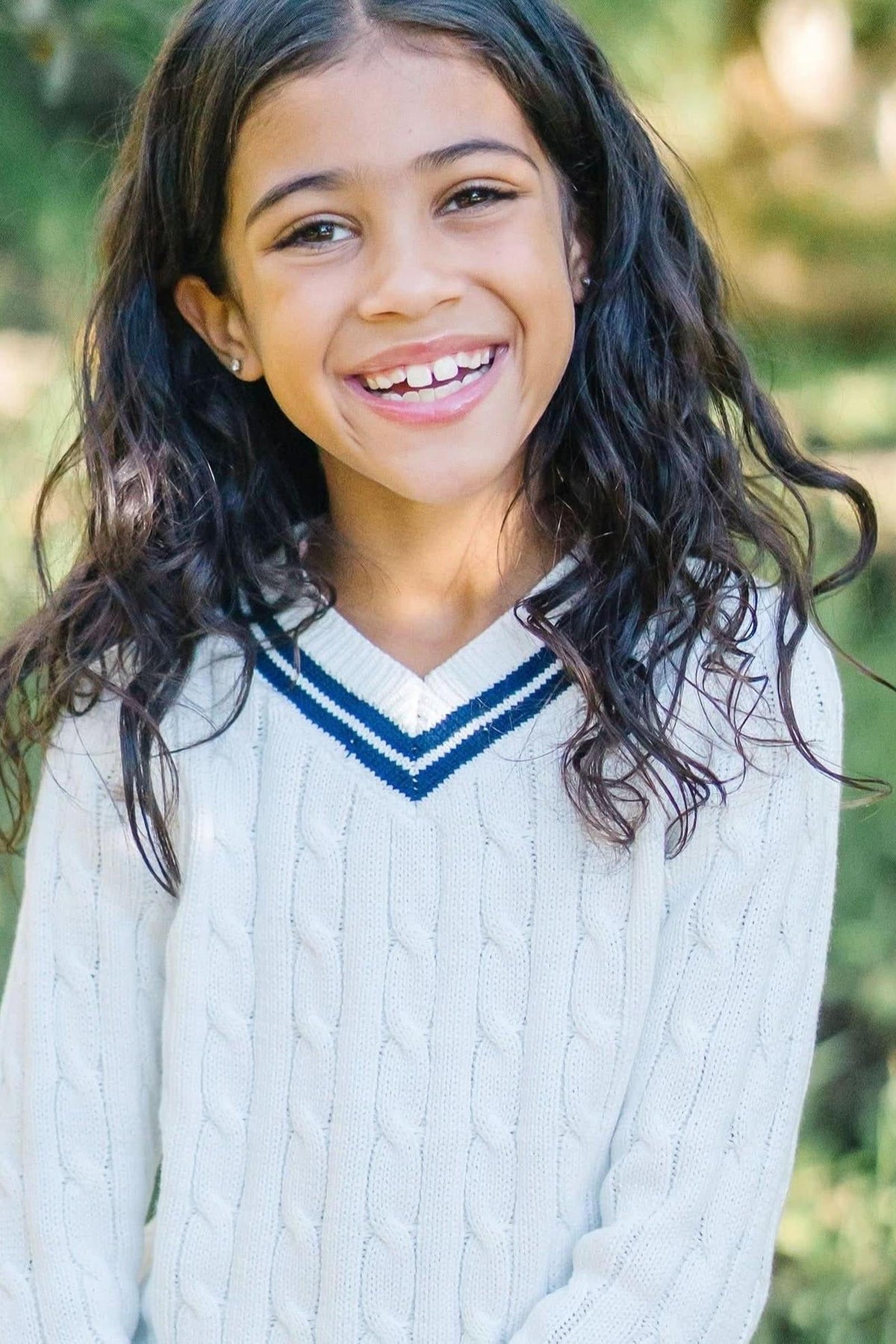 A smiling girl with long, curly hair wears a white cable-knit sweater featuring a blue-striped v-neck.