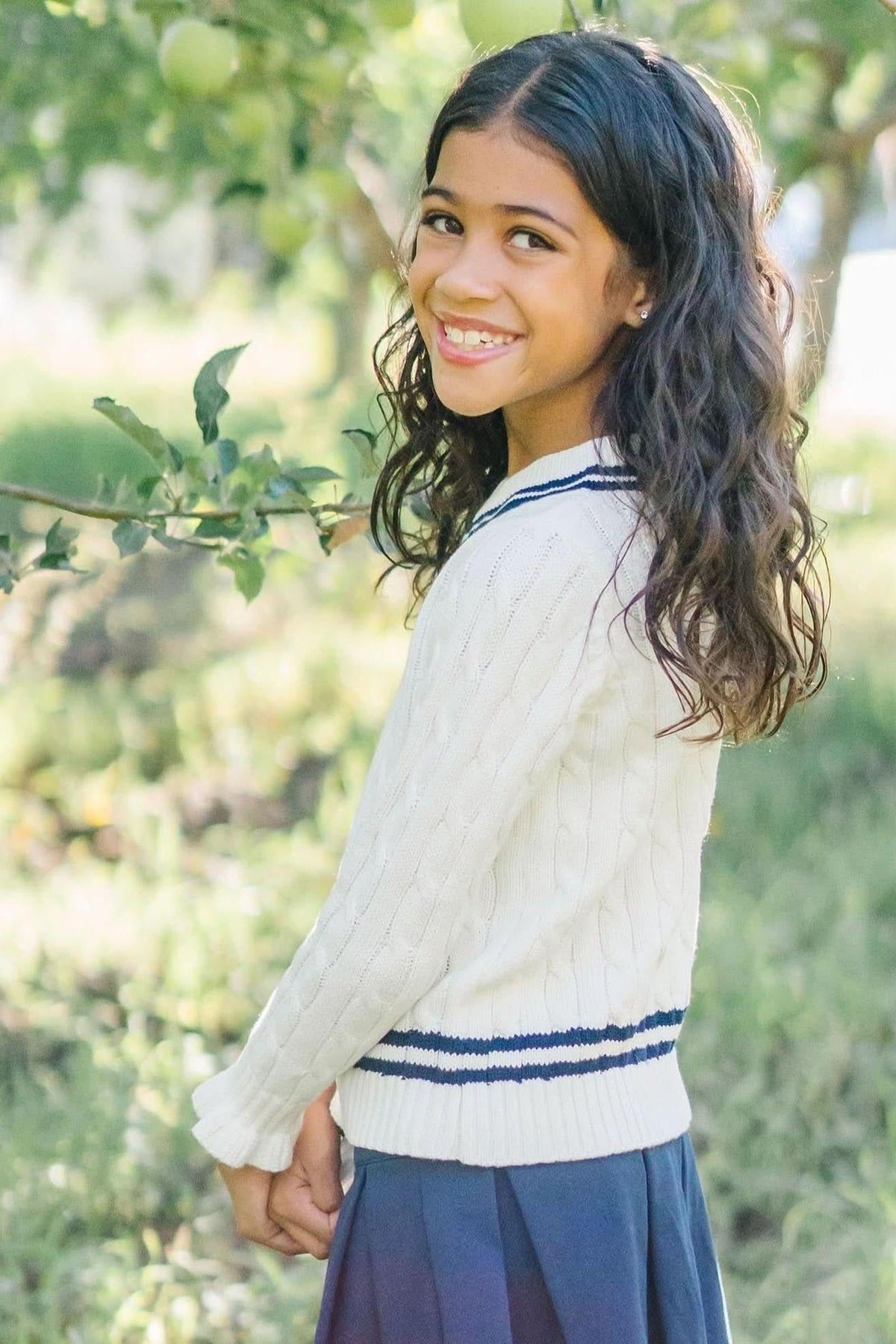 A smiling girl with long, curly hair wears a white cable-knit sweater with navy stripes, standing in a lush, green.