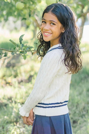 A smiling girl with long, curly hair wears a white cable-knit sweater with navy stripes, standing in a lush, green.