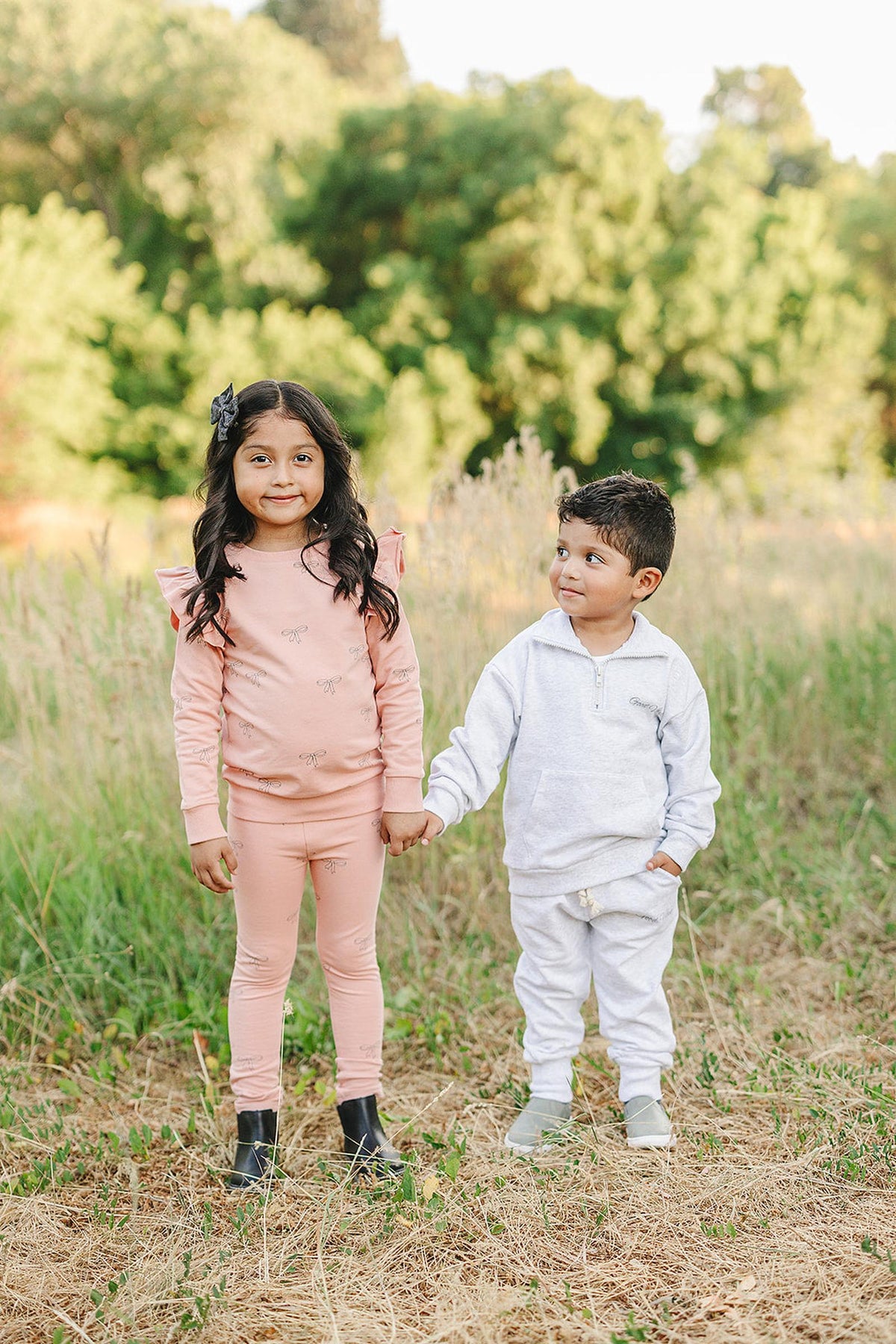 A girl in a pink outfit and a boy in a grey quarter-zip sweatshirt stand holding hands outdoors.