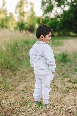 A child wearing a grey Good Vibes Club sweatshirt and pants stands outdoors in a grassy area.