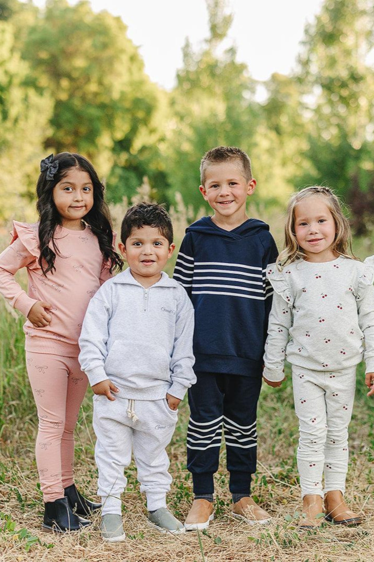 Four children in colorful casual outfits stand outdoors on grass, smiling against a backdrop of green trees.
