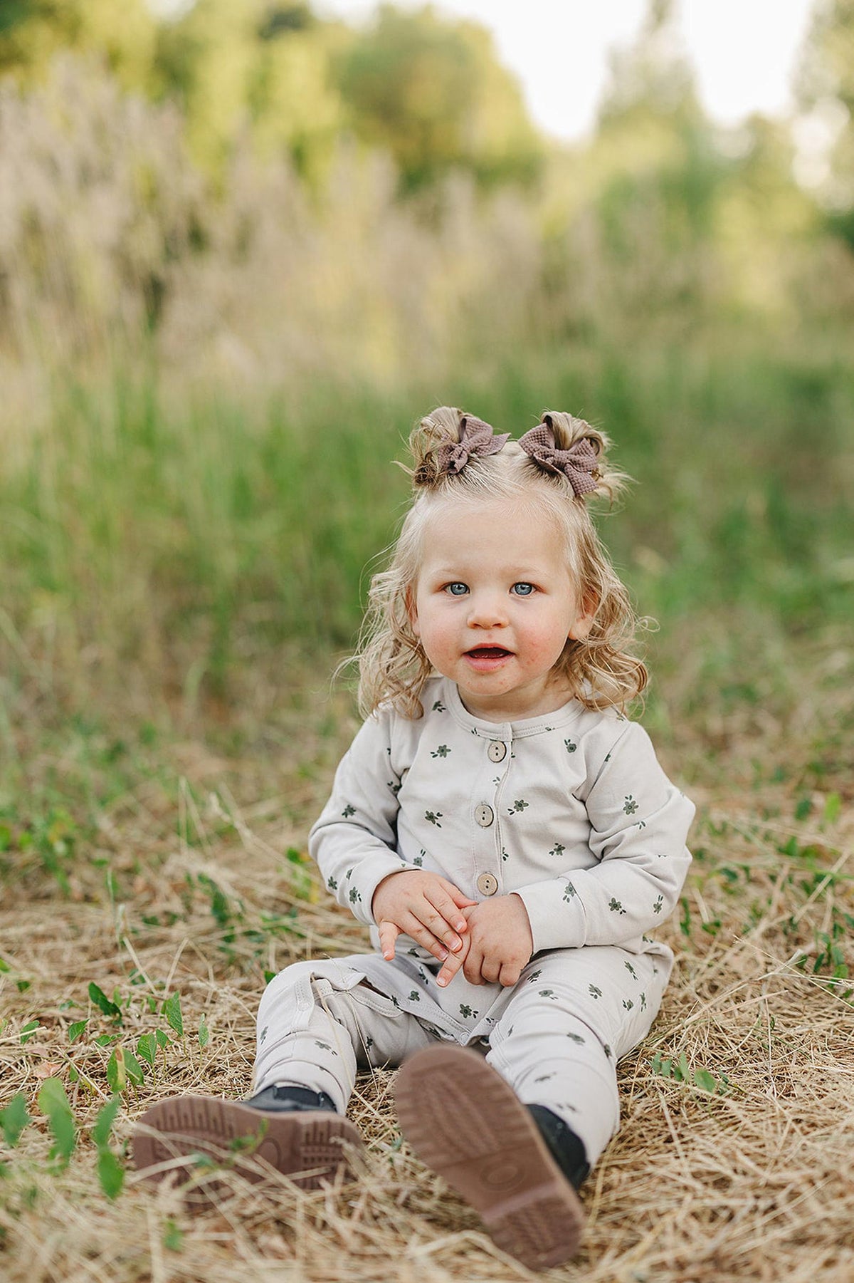 A toddler in a floral-patterned romper sits on grass with greenery in the background, wearing hair bows.