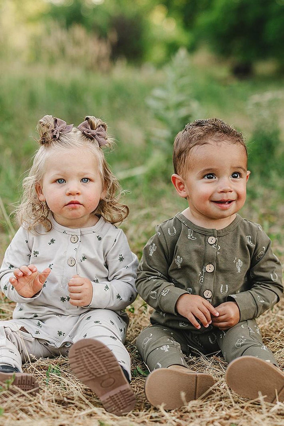 Two toddlers sit outdoors wearing button-up rompers with western patterns, one in light gray and one in olive green.