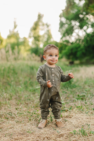A toddler wearing a green, western-patterned romper with buttons, standing outdoors on grass with trees in the background.