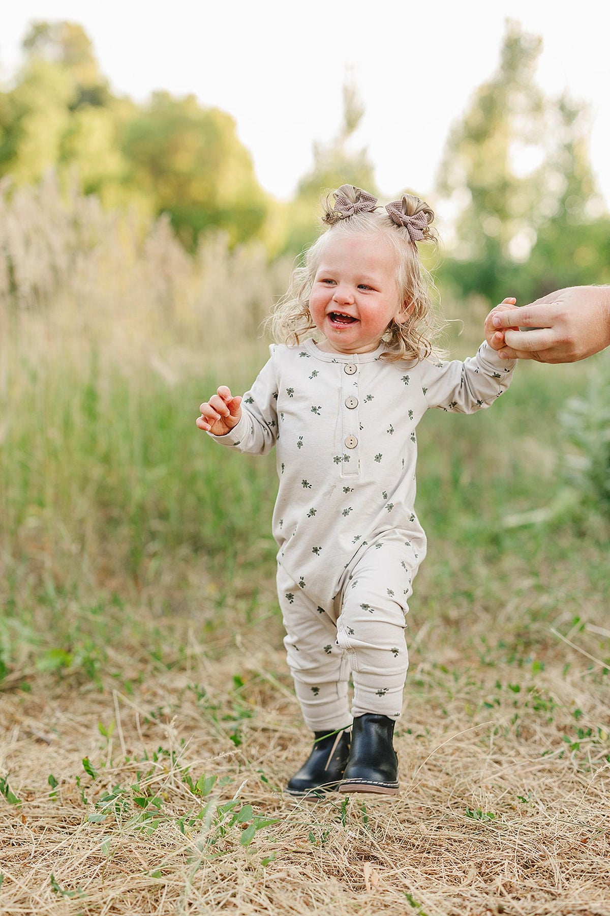 A smiling toddler in a floral romper and black boots walks outdoors, holding an adults hand.