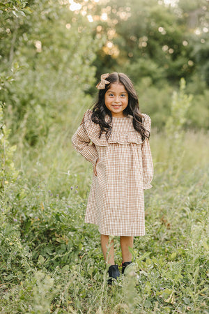 A young girl stands in a grassy field, wearing a gingham ruffle long sleeve cotton dress in tan and cream. She smiles and has a matching bow in her hair.