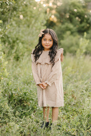 A young girl stands in a grassy field wearing a gingham ruffle long sleeve dress in warm tones. She smiles, accessorized with a matching bow in her hair.