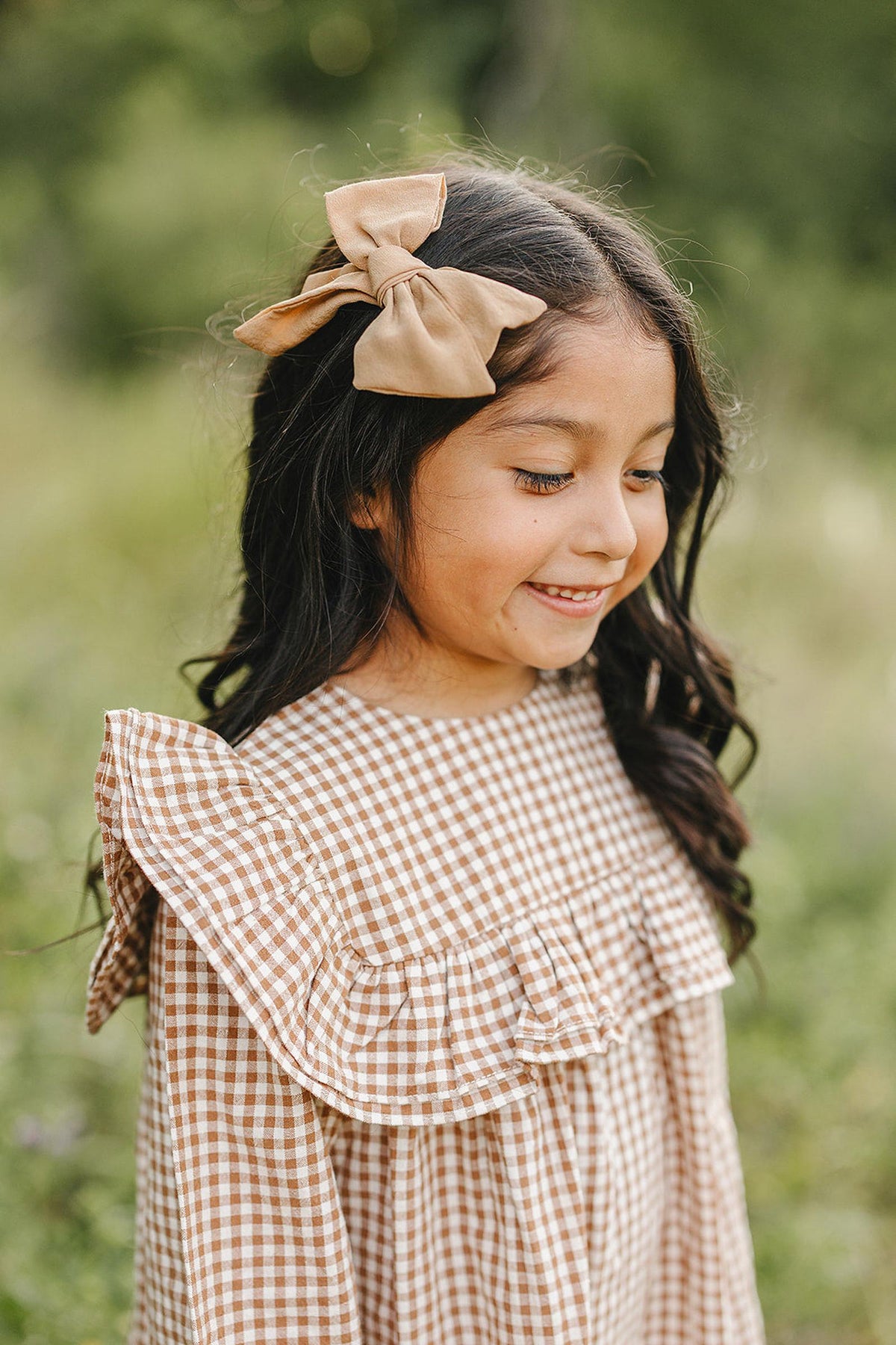 A smiling girl with long, dark hair wears a brown gingham ruffle dress and a matching bow. She stands outdoors, surrounded by greenery, embodying a joyful, playful spirit.