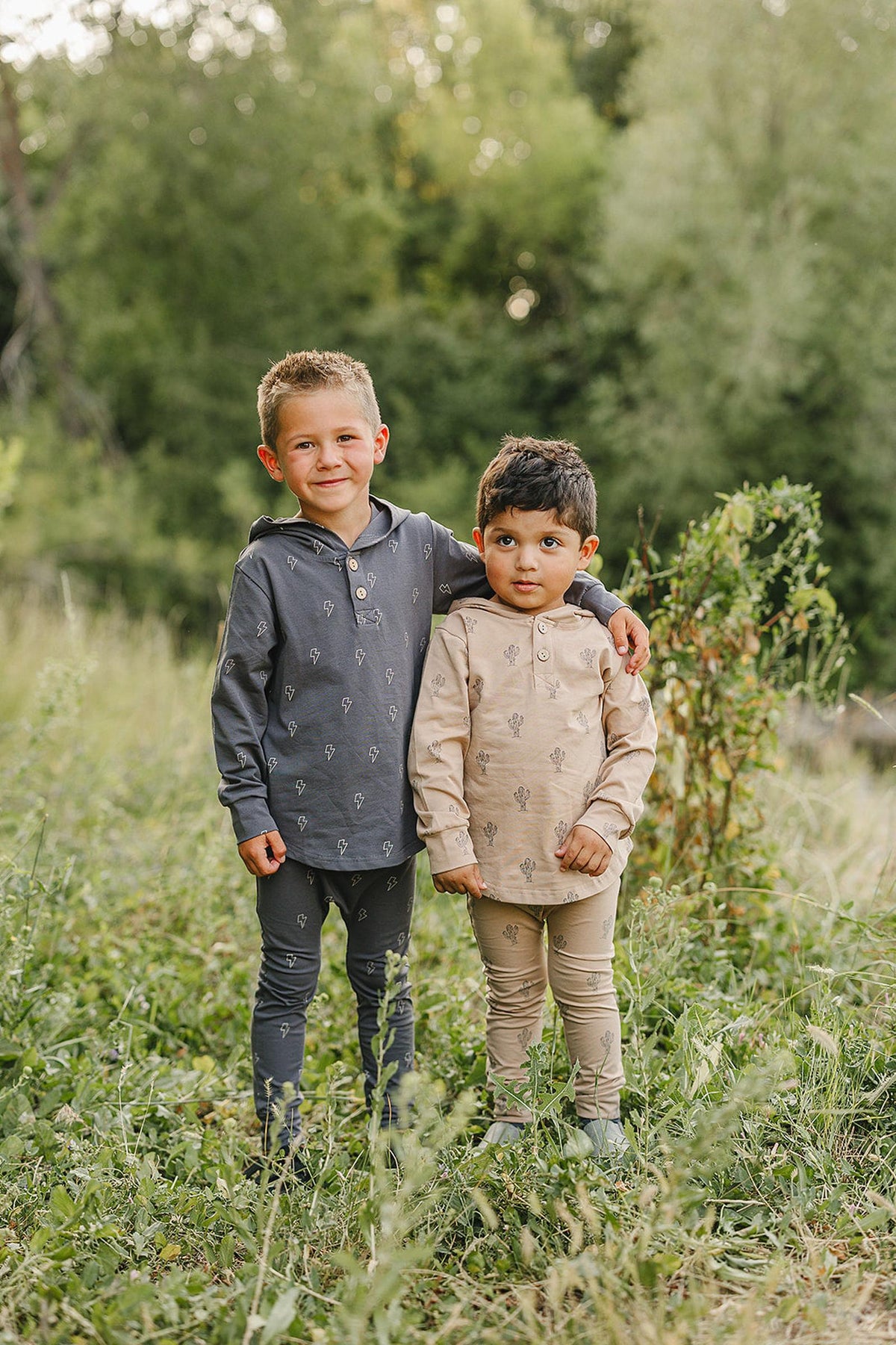 Two children outdoors in matching cactus-patterned hooded sets, one in gray and one in beige, standing on grass.